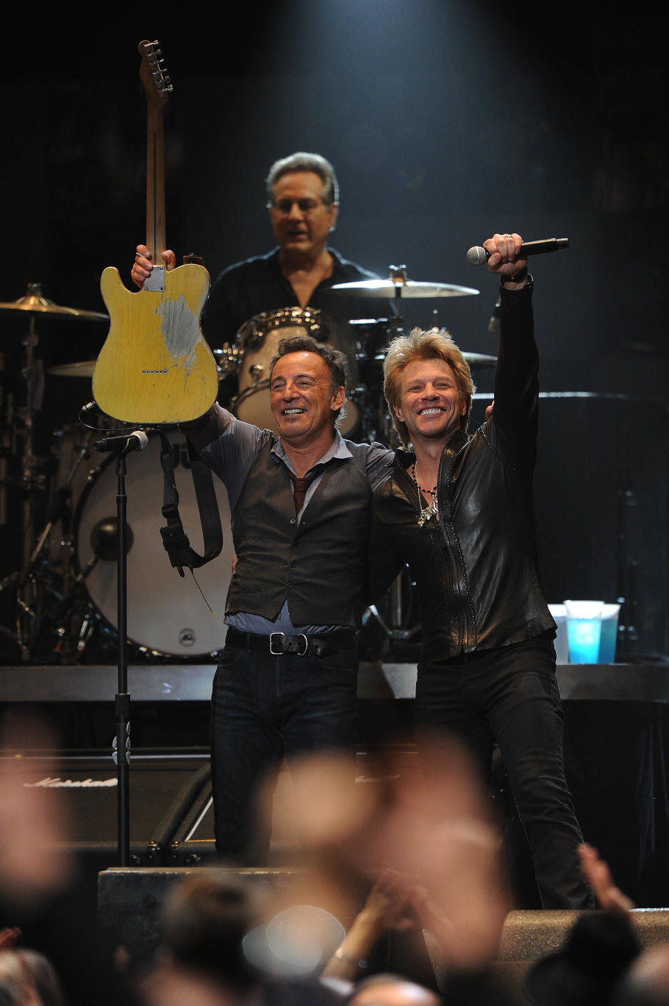 Bruce Springsteen, left, and Jon Bon Jovi onstage together, with Springsteen raising his guitar in the air and Bon Jovi raising the microphone in the air.