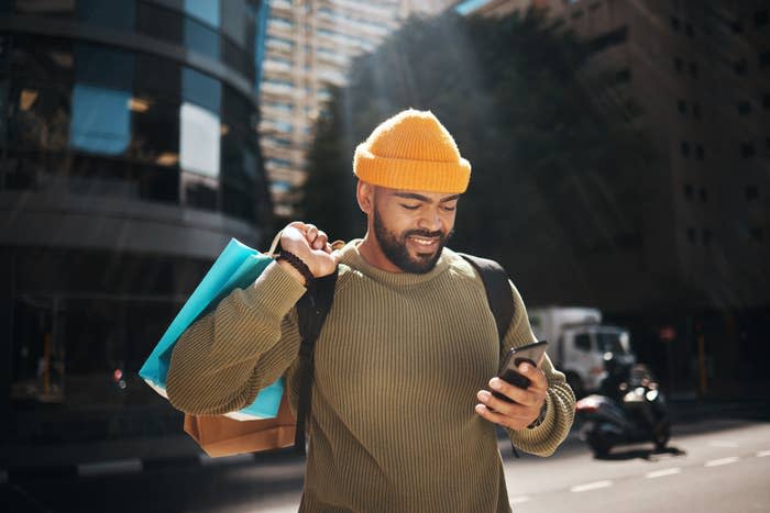 Man with a beard wears a yellow beanie and green sweater, smiles at phone while holding shopping bags on city street