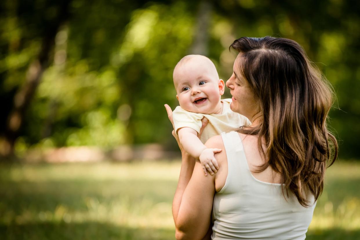 Middle-aged mother back facing the camera, holding her baby up, baby is facing the camera looking off to the left, with a blurred background of grass and trees in a park