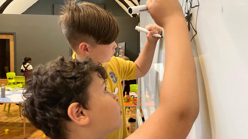 Two students in a classroom draw on a whiteboard.  The boy closest to the camera has curly brown hair and is drawing a spider with a black marker.  Your classmate has partially shaved hair and is drawing something with green paint.