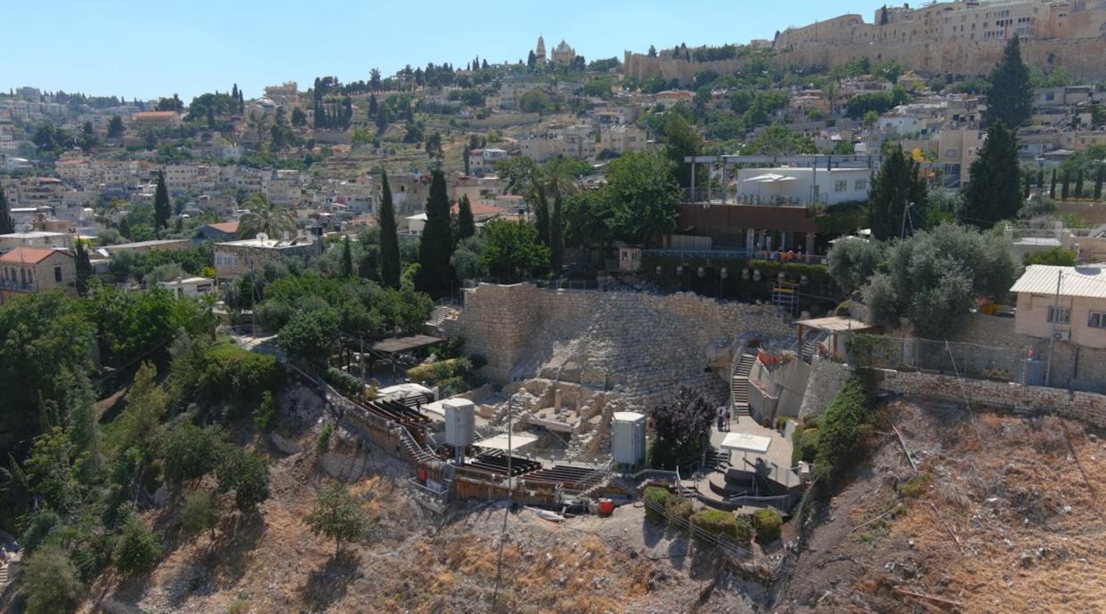  Zoomed out view of a stone structure at an archeological site with other buildings surrounding it. Trees are growing around the site and between the city buildings. 