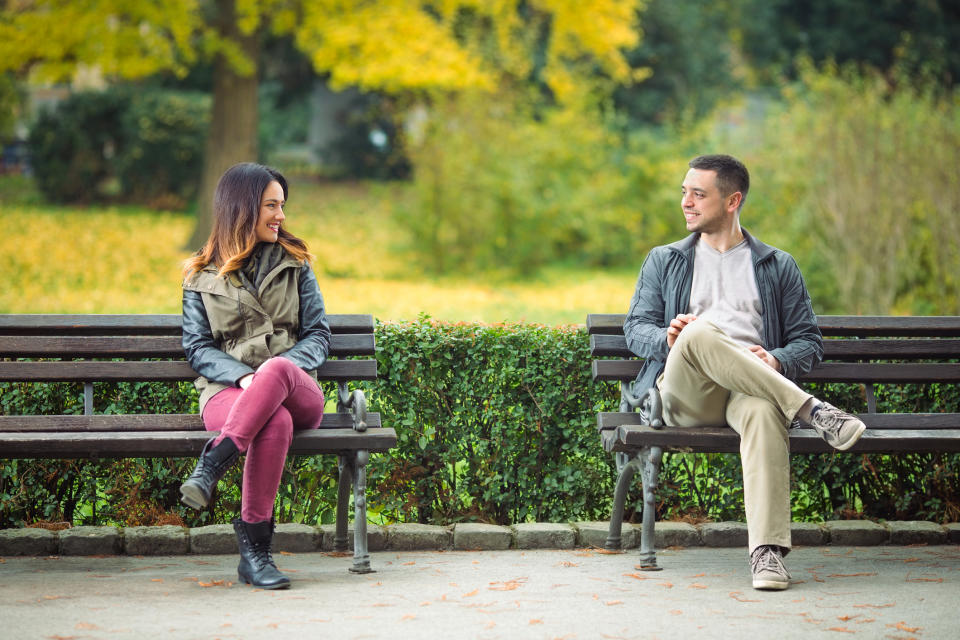 A woman and man sit on two separate benches in a park, smiling at each other