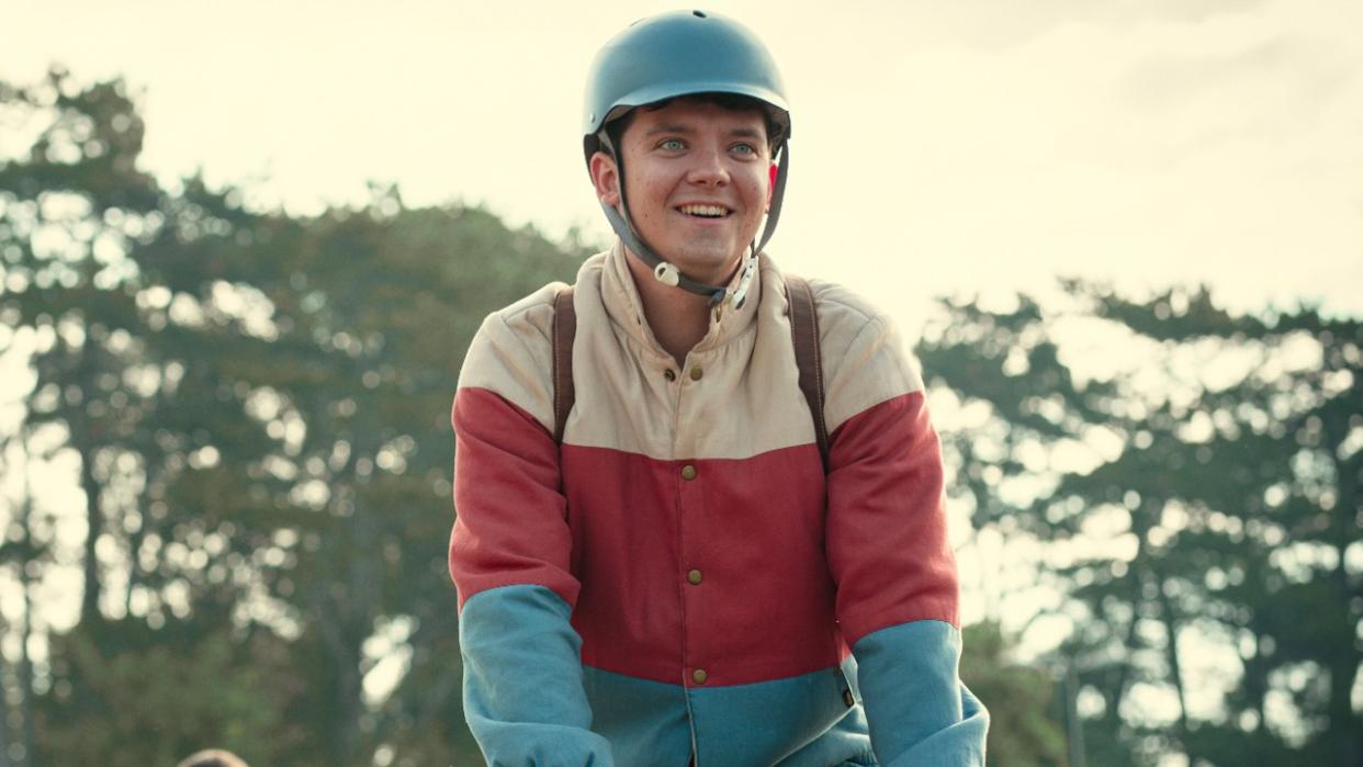  A press photo from Netflix of Asa Butterfield as Otis riding a bike wearing his white, red and blue coat in Season 4 of Sex Education. 