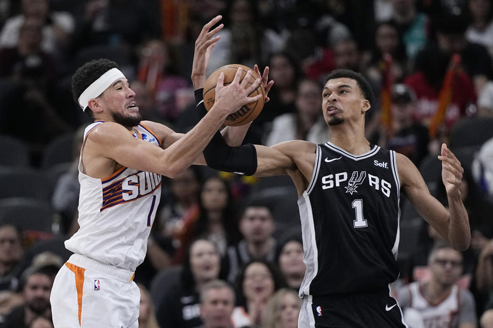 Phoenix Suns guard Devin Booker, left, and San Antonio Spurs center Victor Wembanyama, right, reach for a rebound during the second half of an NBA basketball game in San Antonio, Saturday, March 23, 2024. (AP Photo/Eric Gay)