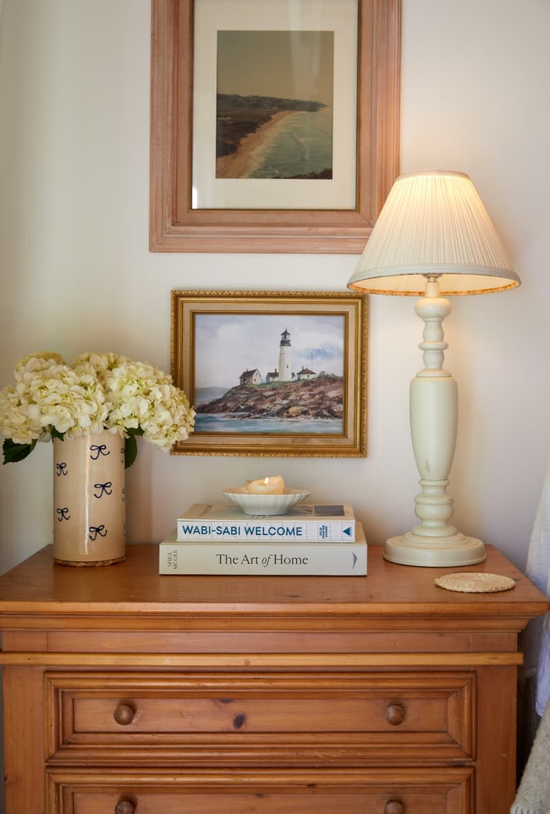 A bedside table with hardcover books, a vase of flowers, and a lamp.