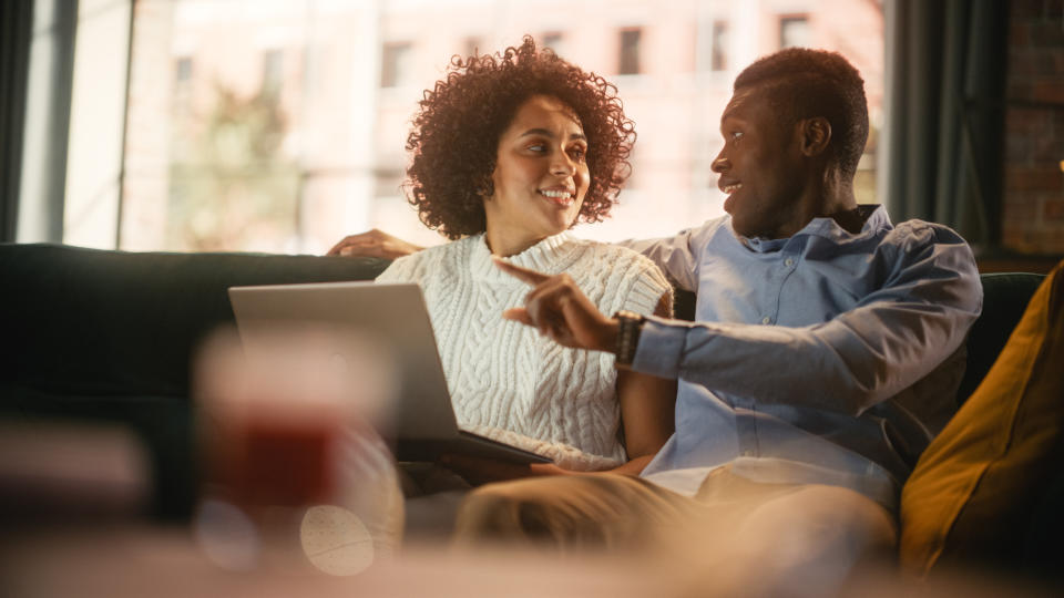 Multiethnic Couple is Sitting on Couch in Stylish Living Room and Choosing Items to Buy Online with Laptop Computer. Black Man Embracing His Wife with Love and Support. Black Family Business.