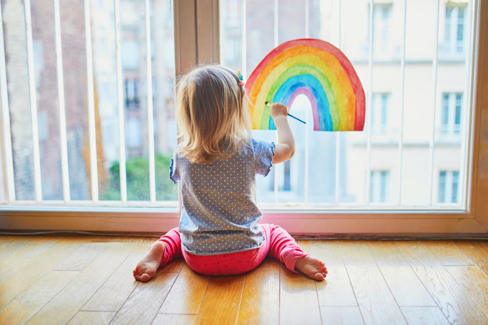 Adorable toddler girl painting rainbow on the window glass as sign of hope. Creative games for kids staying at home during lockdown. Self isolation and coronavirus quarantine concept