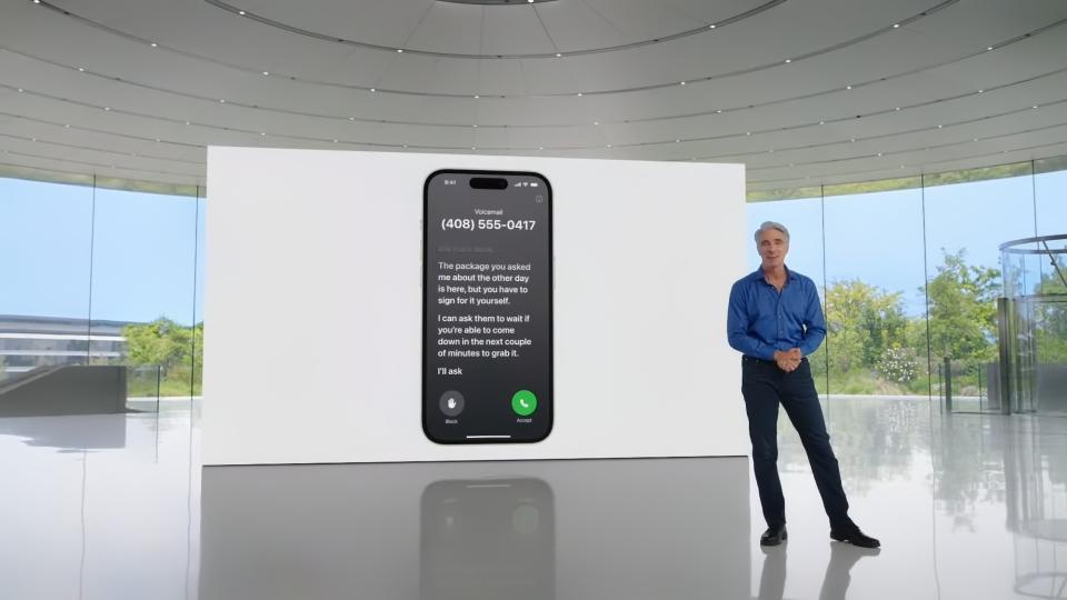 Apple executive Craig Federighi (blue button-down shirt, black slacks) standing in a room in Apple Park (modern gray aesthetic with trees visible outside the windows). On a screen behind him is a demonstration of live voicemail.