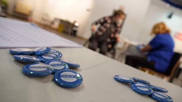 A pile of  'I got COVID-19 vaccination' pins on a table at the Centre square Mall COVID-19 vaccination clinic in Yellowknife. In the background, Riley Oldford receives his first dose of the COVID-19 vaccine on Thursday.