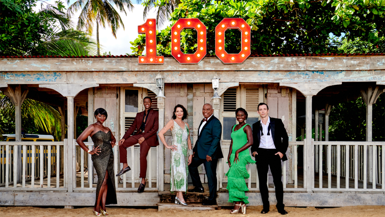  Death in Paradise season 13 cast pose outside the shack with a 100 sign on top. 