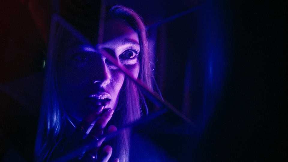 a blonde woman looks into a broken mirror in a purple-tinged room