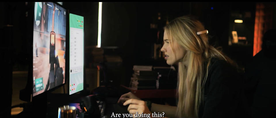 Sasha Luss interacts with computer screens on a set, subtitle reads "Are you doing this?"