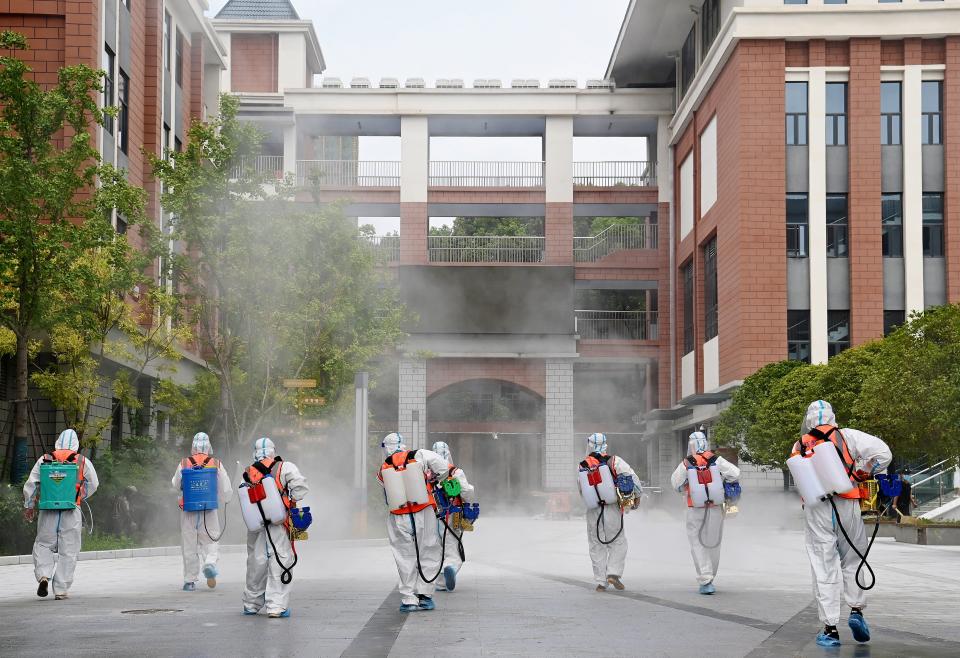 Eight workers in protective suits disinfect the pavement in front of a school.