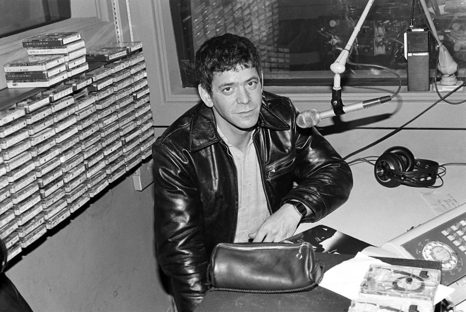 lou reed sits at a table with a microphone hanging in front of him, he looks at the camera and wears a leather jacket and shirt