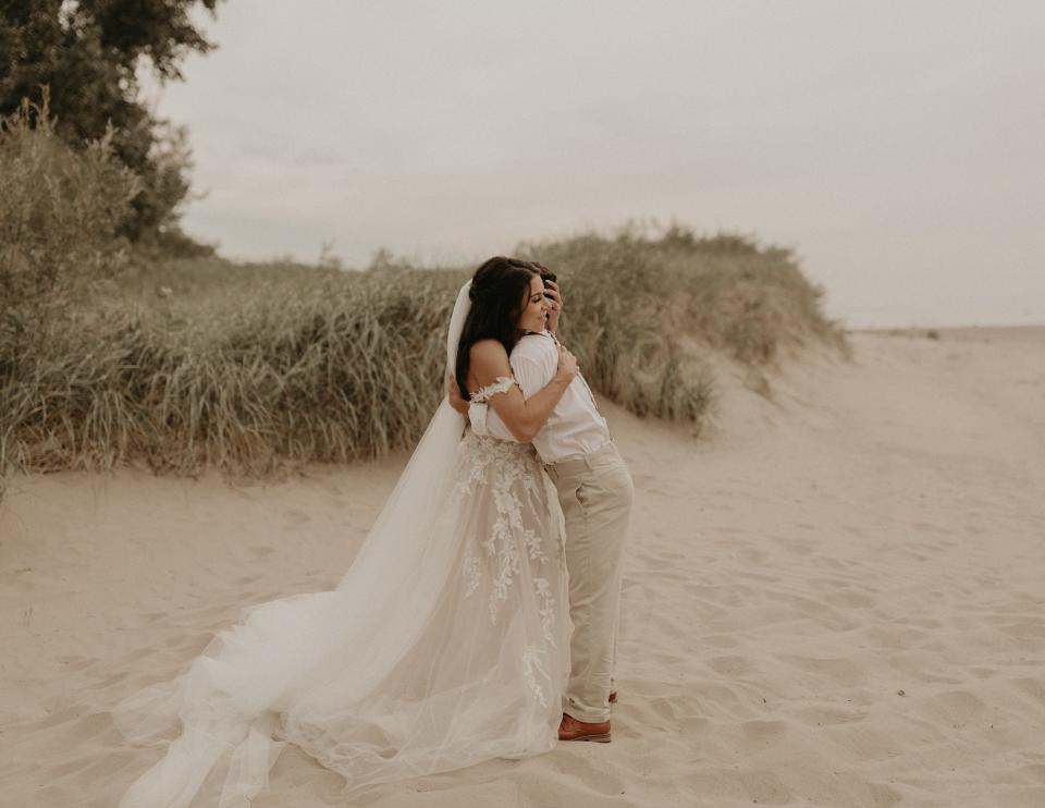 A bride and her brother hug on a beach.