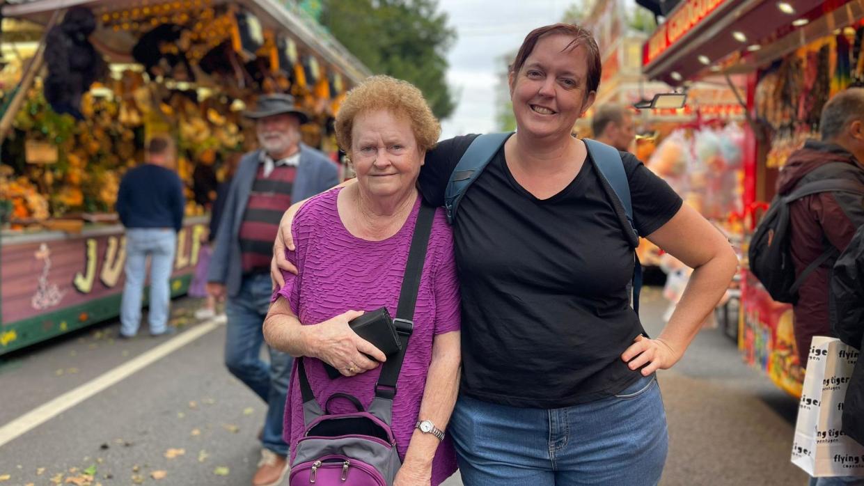 Two women, one a generation older than the other, cuddle and pose for a picture on the fairground path.