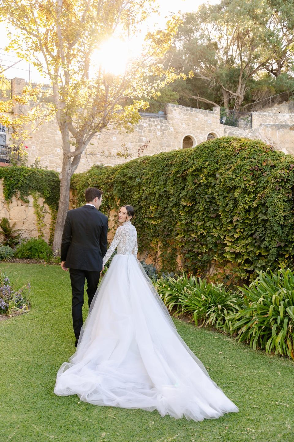 A bride looks over her shoulder as she and her groom hold hands and walk away from the camera.