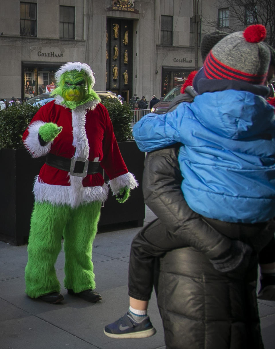 In this Friday, Dec. 20, 2019 photo, a costumed Grinch performer, left, works a sidewalk for tips along 5th Avenue near Rockefeller Center in New York. Some performers, who solicit tips from tourists from designated "activity zones" in Times Square, have been migrating to Rockefeller Center for the holiday season where no such zones exist. (AP Photo/Bebeto Matthews)