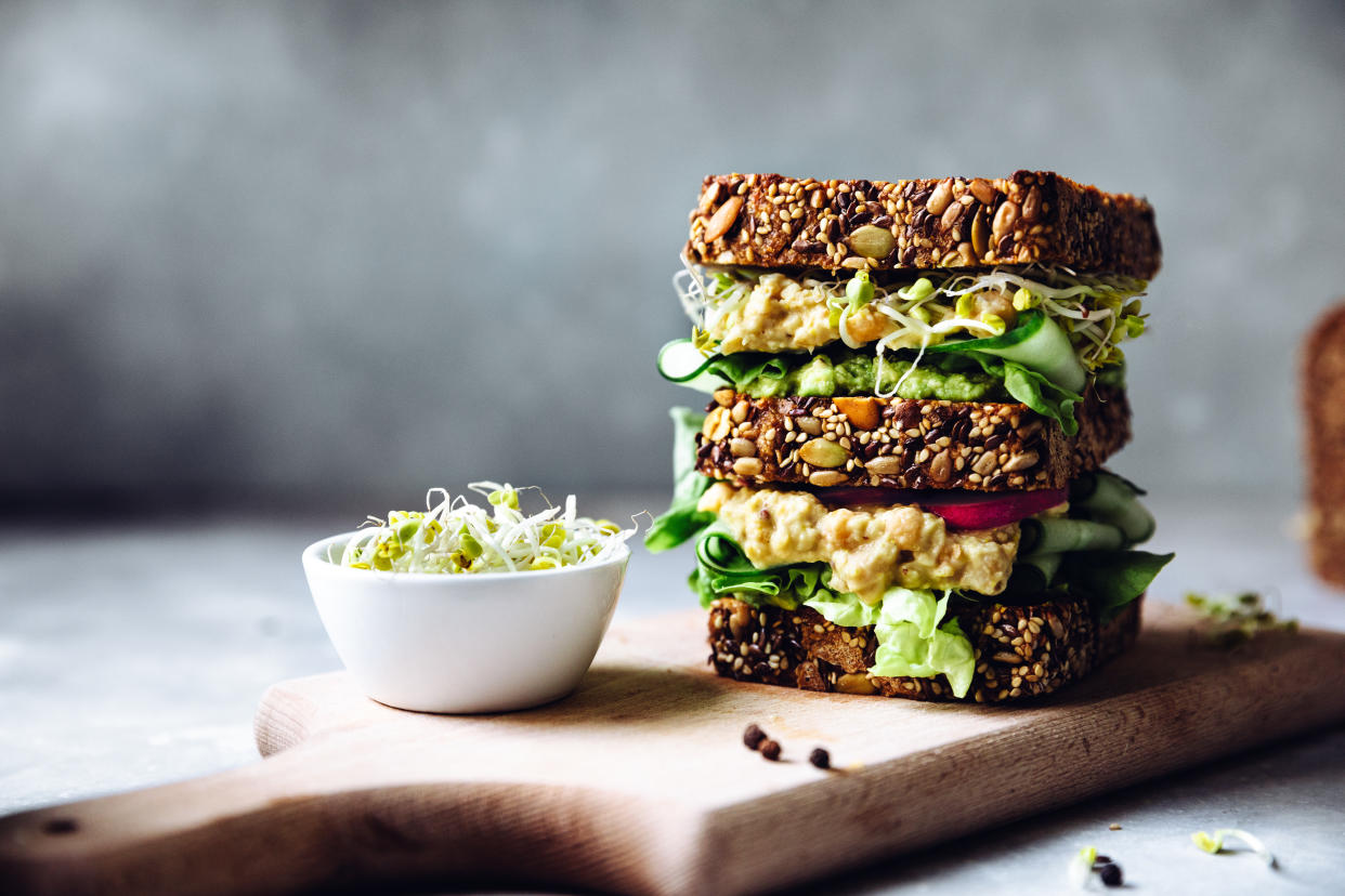 Vegetarian sandwich made with sourdough bread, avocado creme, cucumber, radish and remoulade sauce with bowl of sprouts served on a table.