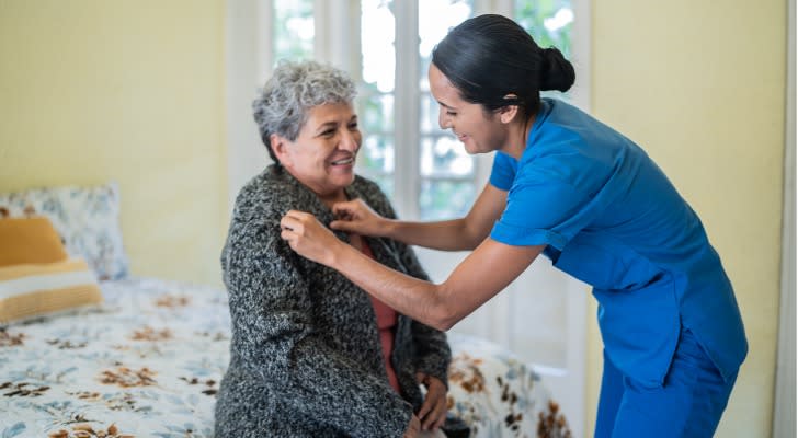 A nursing home employee helps a resident put on a sweater inside her room. 