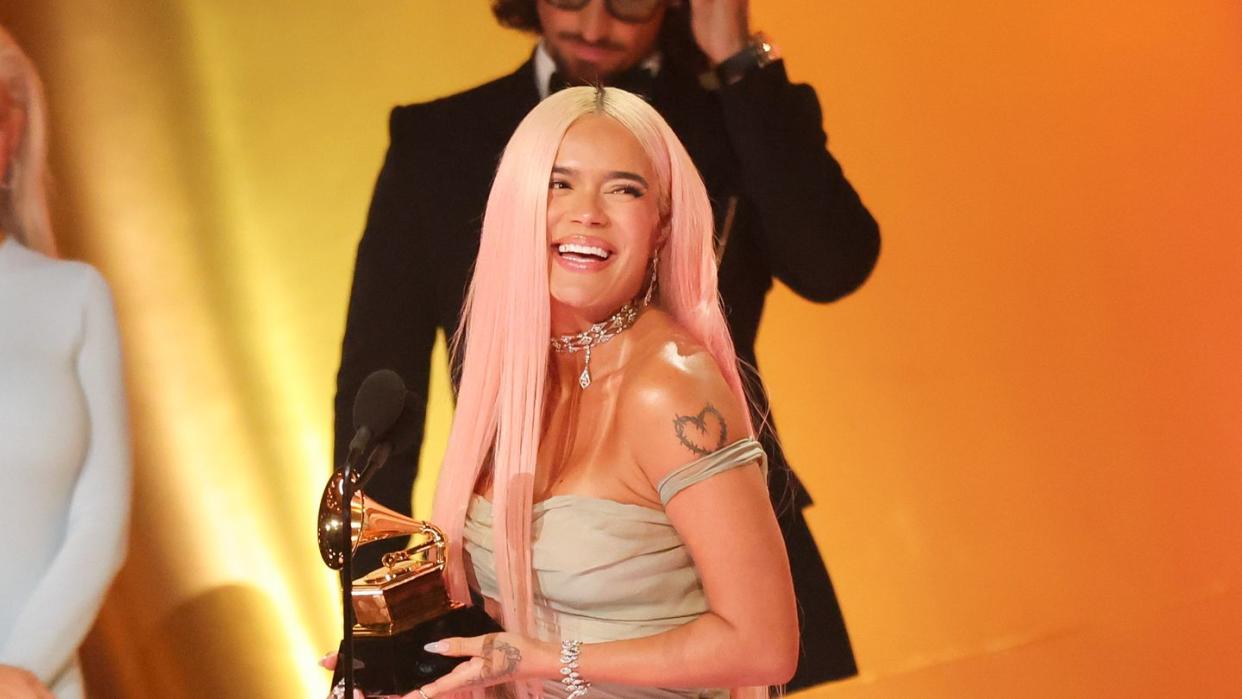 karol g smiles and looks to the right, she holds a grammy trophy and stands behind a microphone in a strapless gray dress