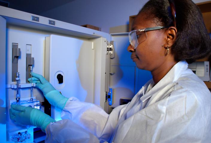 Laboratory worker Karidia Diallo, wearing gloves, a lab coat and protective eyewear, places samples into a DNA analyzer machine.