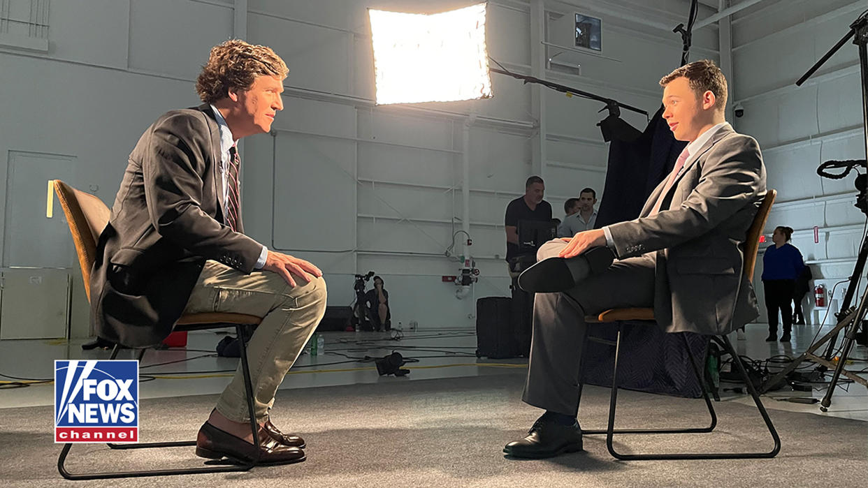 Kyle Rittenhouse sits in a large room under boom-mounted lights facing Tucker Carlson. A Fox News logo is in a corner of the frame. (Courtesy of Fox News via AP)