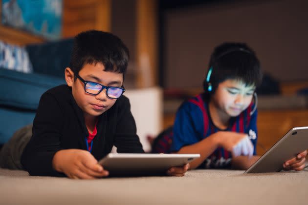 Two multiracial kids at home lying on carpet with devices in their hands playing games.