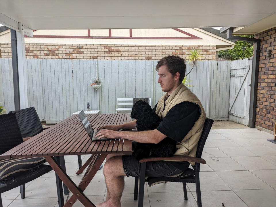 Man sits at outside table with dog on lap and a laptop.