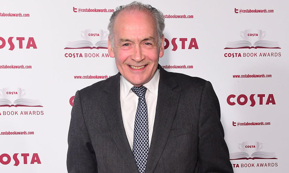 Alastair Stewart, wearing a black suit jacket over a white shirt and patterned tie, smiles as he attends the Costa Book Awards