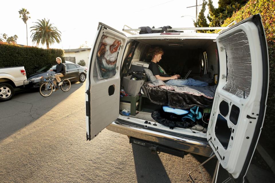 A person works on a laptop in the back of van with its back doors opened.