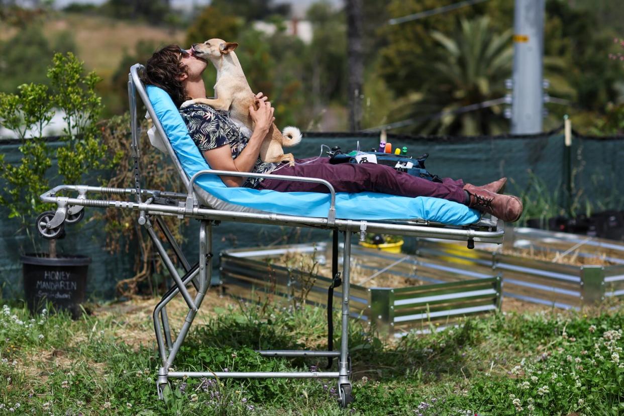 A man kisses a dog while lying on a gurney in a yard.