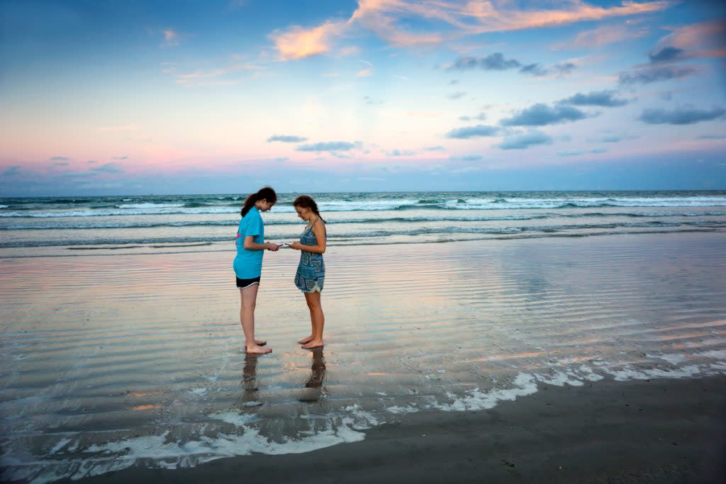 Two teenage girls look at their phone while standing in the water at Coca Beach during sunset. 