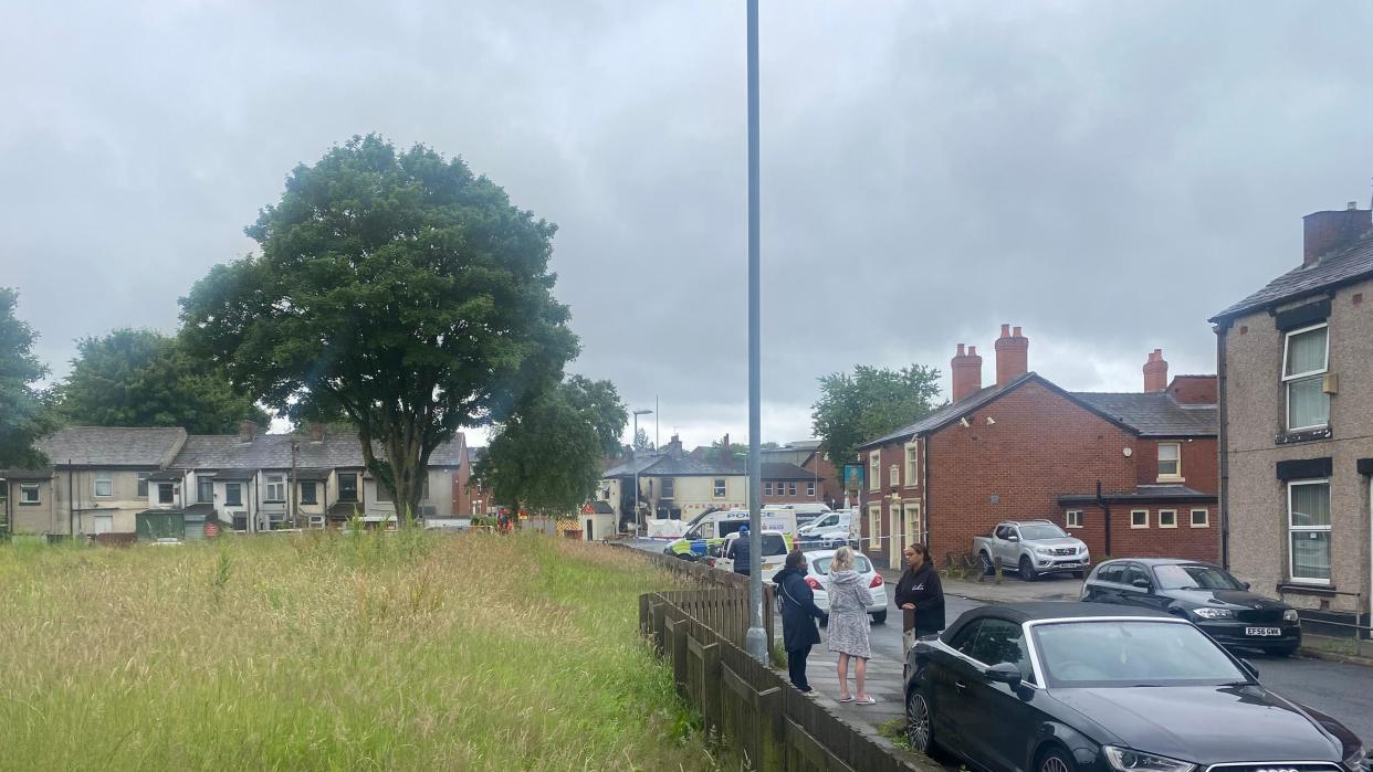 People gather in front of a police cordon, with a ruined pub visible in the background