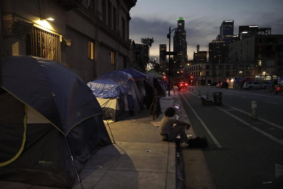 A man sits on a sidewalk as dusk settles over Skid Row in downtown Los Angeles