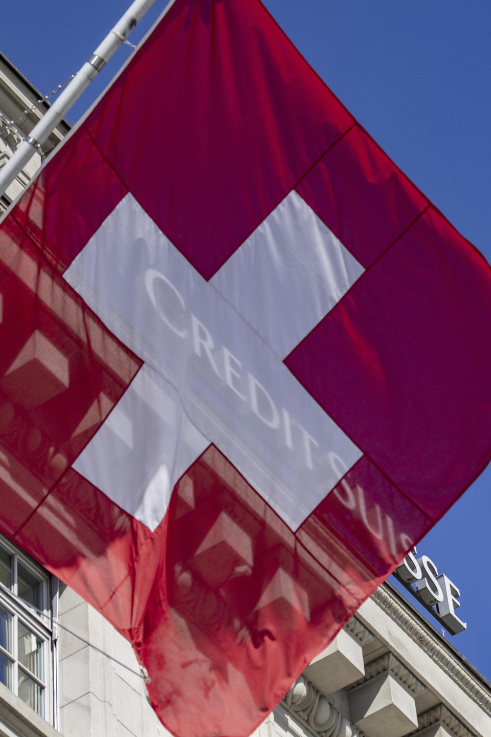 Switzerland's flag flutters in front of the Swiss bank Credit Suisse CS logo in Lucerne, Switzerland, Thursday March 16, 2023. Credit Suisse, which was beset by problems long before the U.S. bank failures, said Thursday that it would exercise an option to borrow up to 50 billion francs ($53.7 billion) from the Swiss National Bank. (Urs Flueeler/Keystone via AP)