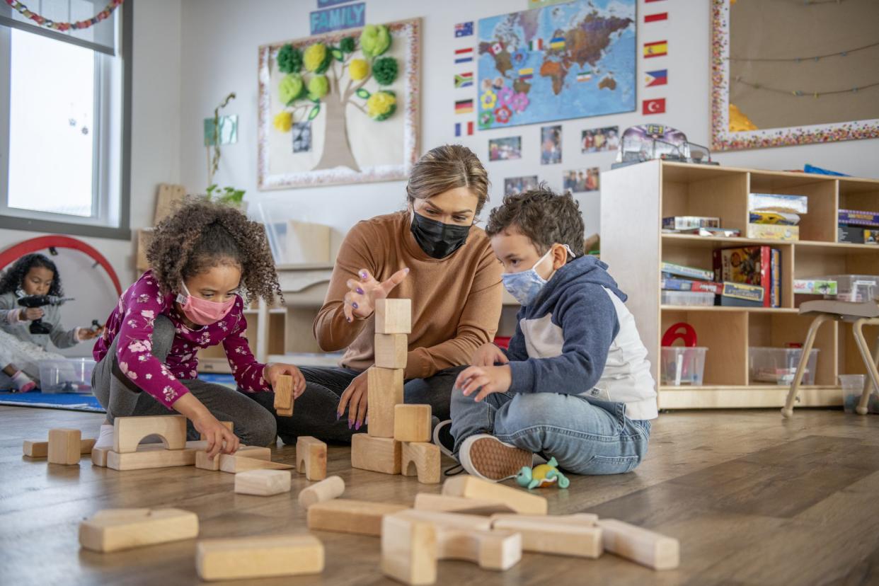 A teacher helps daycare kids build a structure with wooden blocks on the floor.