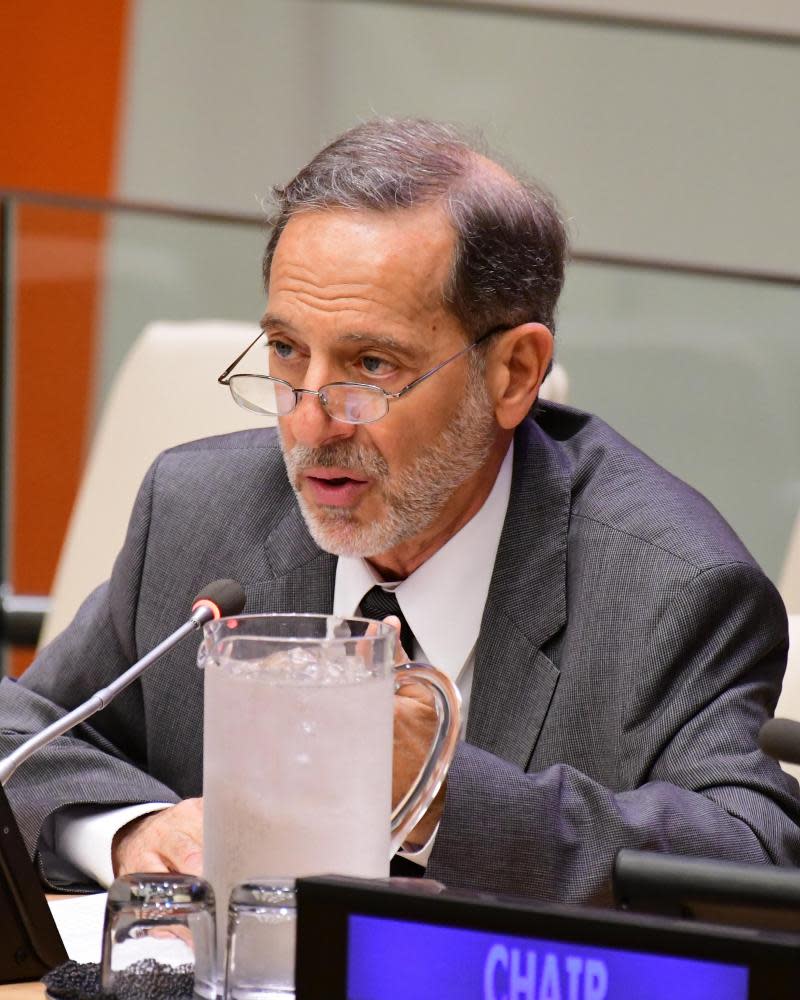 A Middle Eastern man with a trim white beard, dark gray hair and a bald spot, with wire-rimmed glasses, wearing a gray suit, speaks into a microphone at what appears to be a large desk, beside a glass pitcher of ice water.