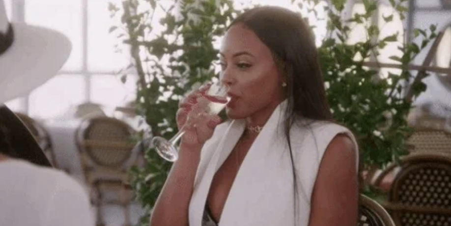 A woman in a sleeveless white top drinks from a glass, sitting outdoors with greenery in the background