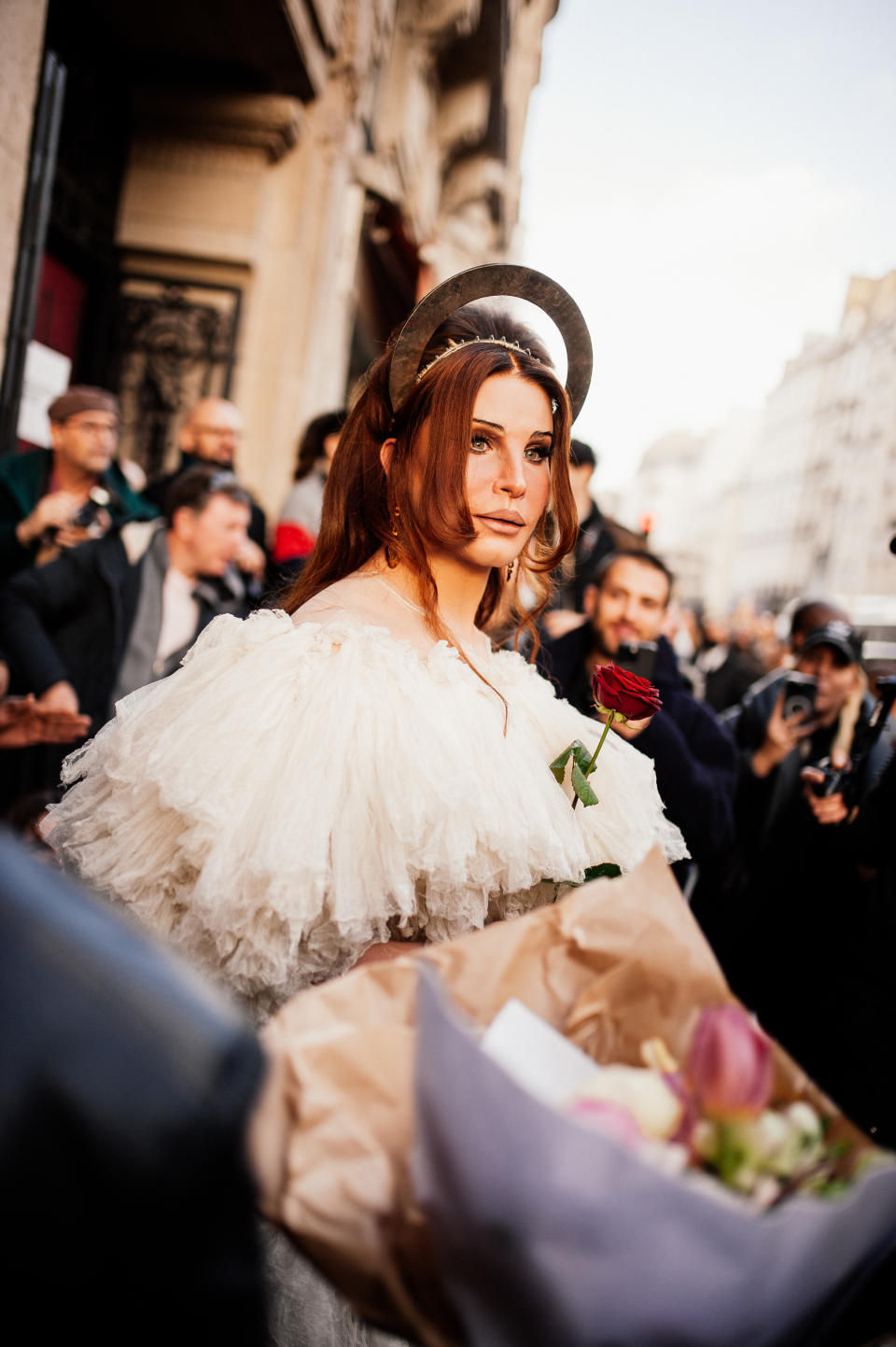 Alexis Stone as Lana Del Rey in a white, fluffy dress holding a red rose, surrounded by photographers outdoors