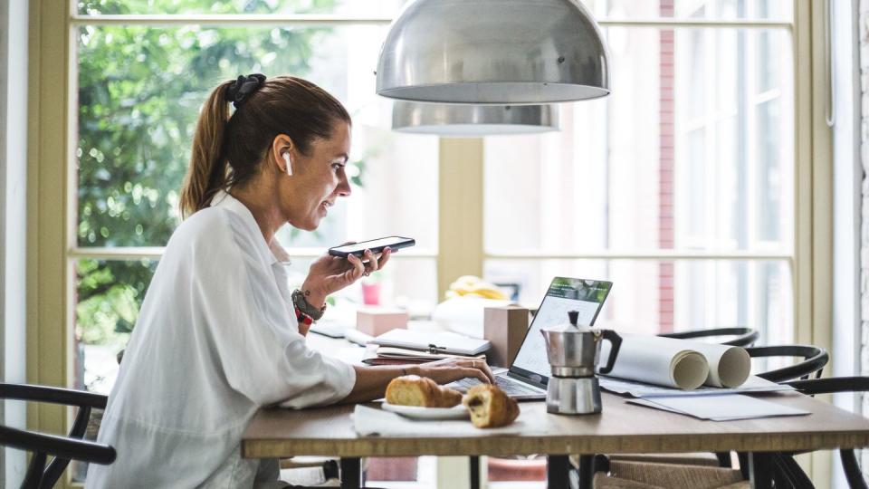 smiling female entrepreneur using laptop while talking through smart phone at home office