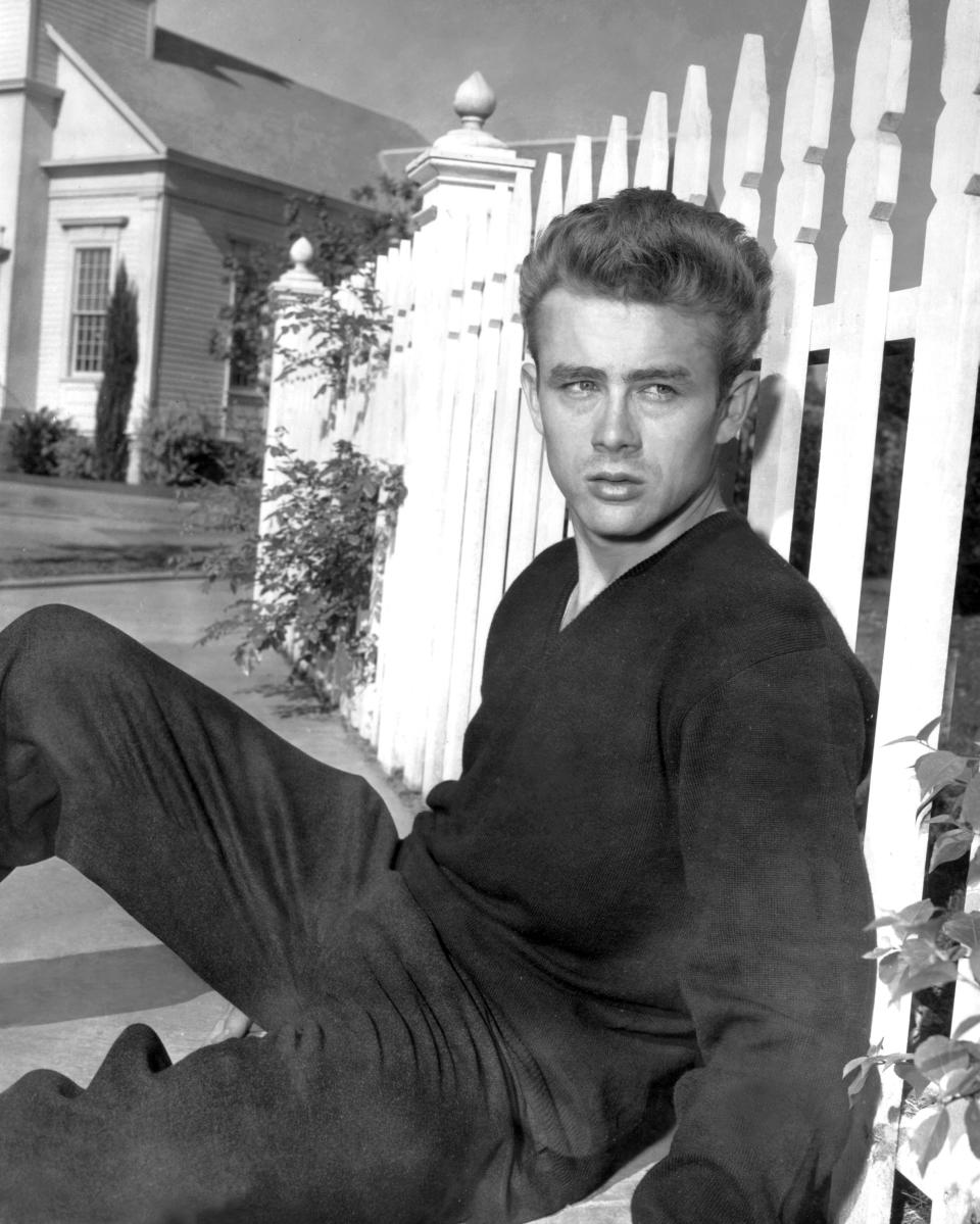 James Dean leans against a white picket fence wearing a V-neck sweater and trousers