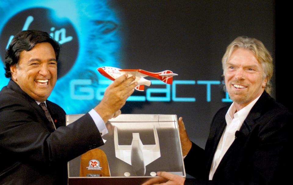 A smiling man with dark hair, left, holds a model of a spaceship next to a bearded man, also smiling, holding a display