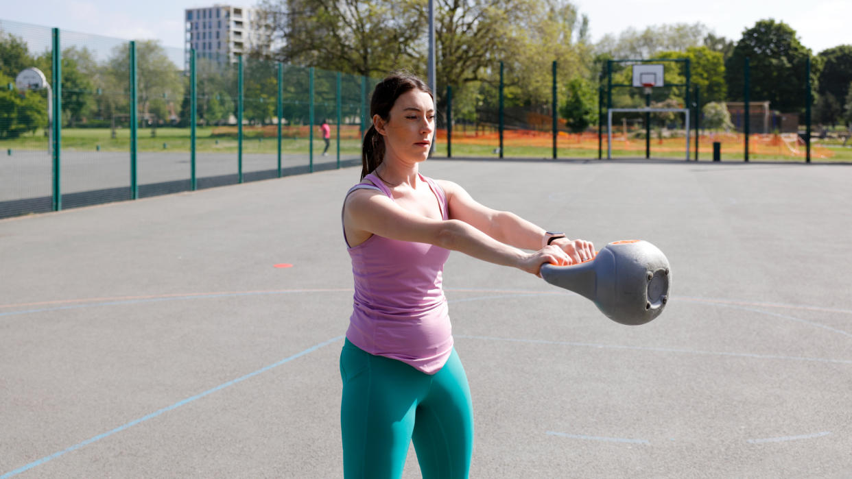 A woman performing a kettlebell swing at the park as part of a kettlebell workout. 