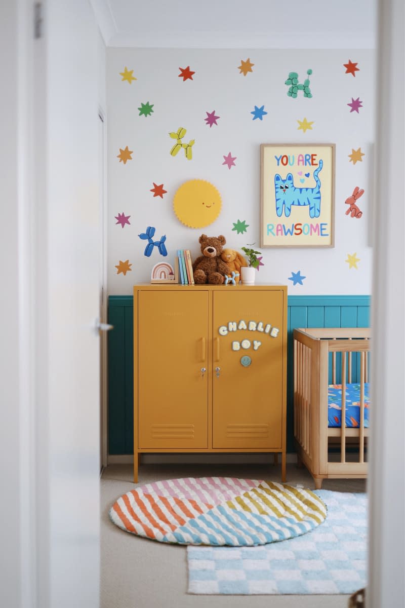 Blue paneling up half wall, balloon dog doodles, yellow locker cabinet next to wood crib, blue and white checkered rug layered with multi colored striped rug
