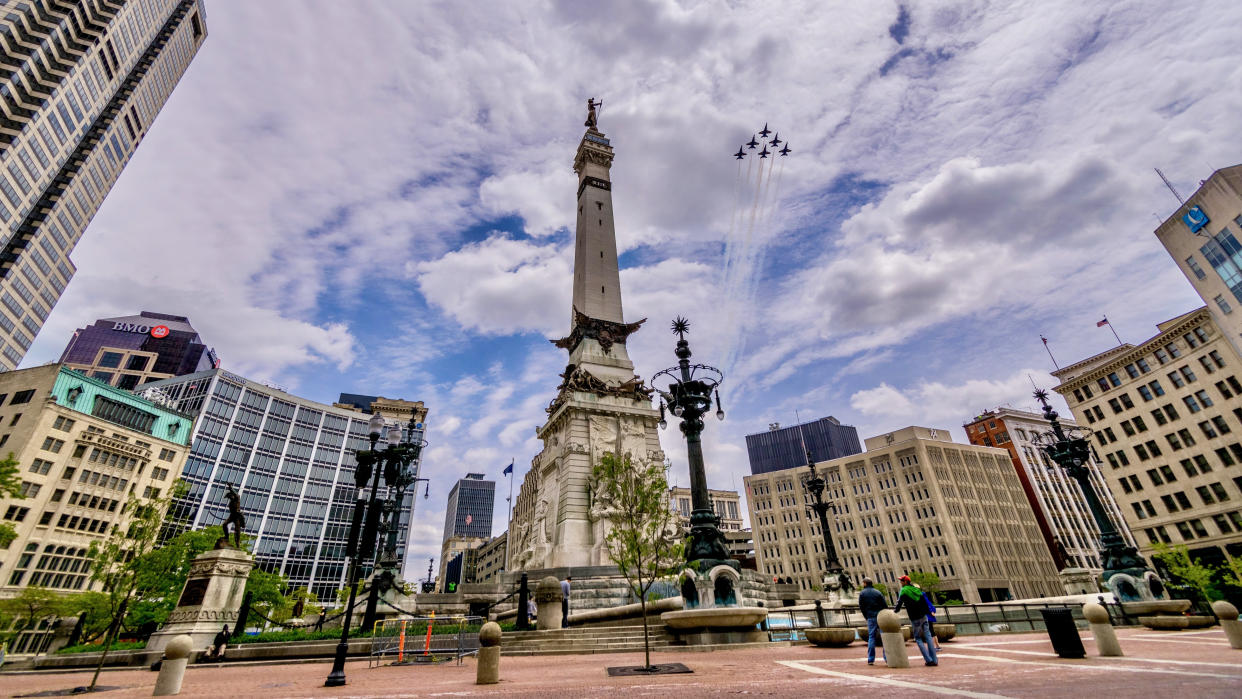  Six jets in formation fly through a cloudy blue sky over a city's center square, a tall monument tower standing in the middle with building surrounding. . 