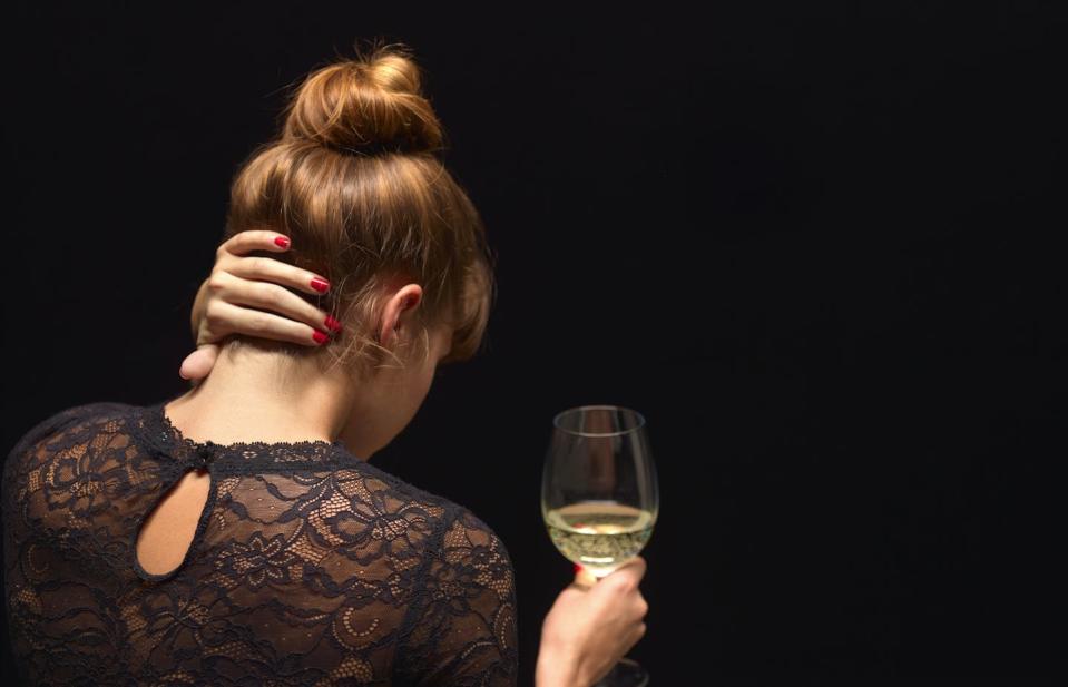 An adult woman looks away from the camera, holds a glass of white wine in one hand and presses her left hand to her neck.