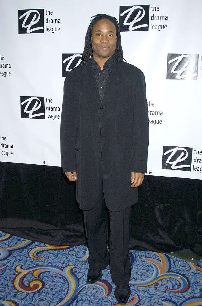 Person in a black suit stands in front of a Drama League backdrop