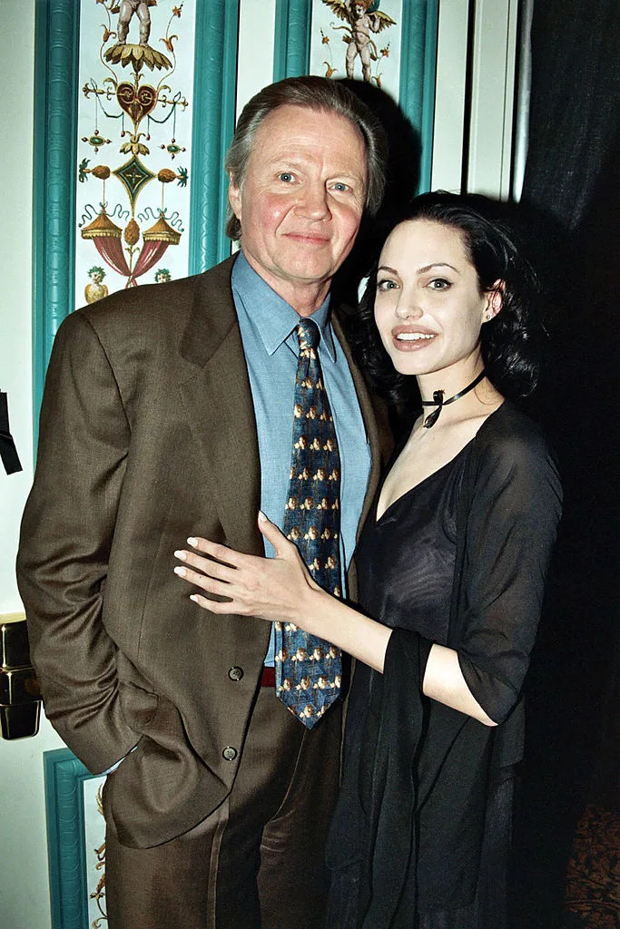 Jon Voight in a brown suit and Angelina Jolie in a black dress with a shawl. They are standing close and smiling for the photo