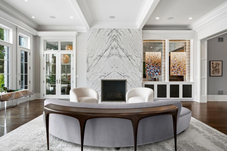 In a light-filled Winnetka home, Inspired Interiors custom-designed the striking marble slab fireplace surround and selected multiple pieces of art. A sofa and curved table by Holly Hunt and a pair of Eichholtz chairs sit atop a Feizy area rug.